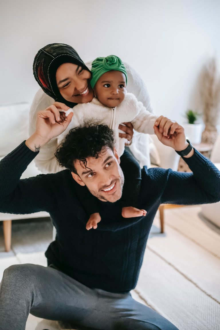 A happy family - the dad sits on the floor while the mom holds the child on dad's shoulders.