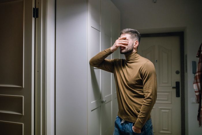 A man wearing a brown sweater stands indoors, appearing distressed with a hand on his face.